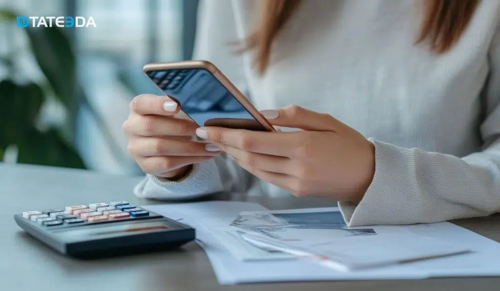 A person using a smartphone while reviewing invoices and calculations, illustrating cost savings with healthcare AP automation by enhancing efficiency, reducing errors, and optimizing payment workflows. | TATEEDA